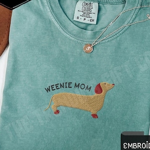 May include: Light green t-shirt with the embroidered text "WEENIE MOM" and a dachshund design. The shirt has a crew neck and short sleeves. The shirt is labeled "Comfort Colors" and is a size small.