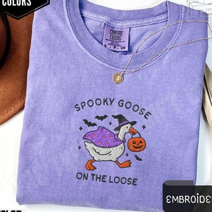 May include: Violet-colored t-shirt with the text "Spooky Goose on the Loose" and an embroidered goose wearing a witch hat and cape, carrying a jack-o'-lantern. The shirt has a Comfort Colors label and a gold necklace.