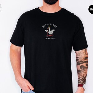 May include: Black t-shirt with a white goose wearing a crown and riding a scooter. The text on the shirt reads "God's Silliest Goose On The Loose".