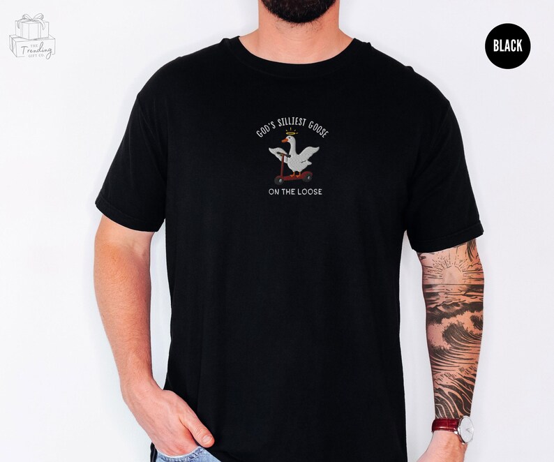 May include: Black t-shirt with a white goose wearing a crown and riding a scooter. The text on the shirt reads "God's Silliest Goose On The Loose".