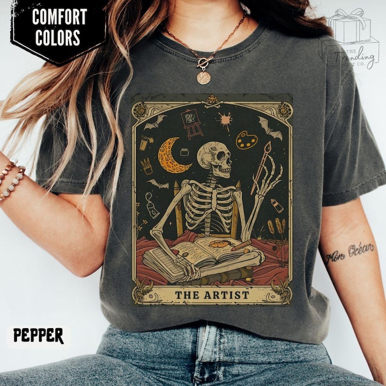 Puede incluir: Camiseta color pimienta con un dise&ntilde;o de carta del tarot con un artista esqueleto. La obra de arte incluye una luna creciente, murci&eacute;lagos, una paleta de pintura y el texto "THE ARTIST". La camiseta tambi&eacute;n tiene el texto "COMFORT COLORS" y "PEPPER".