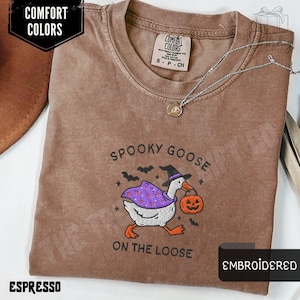 May include: Espresso-colored t-shirt with an embroidered design featuring a goose in a witch hat carrying a pumpkin. The text "SPOOKY GOOSE ON THE LOOSE" is above and below the goose. The shirt has a Comfort Colors label and a silver necklace.