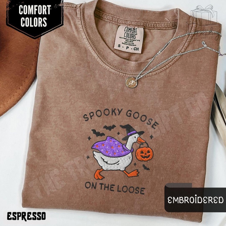May include: Espresso-colored t-shirt with an embroidered design featuring a goose in a witch hat carrying a pumpkin. The text "SPOOKY GOOSE ON THE LOOSE" is above and below the goose. The shirt has a Comfort Colors label and a silver necklace.