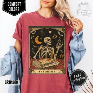 Puede incluir: Camiseta carmes&iacute; con una ilustraci&oacute;n de un artista esqueleto. El dise&ntilde;o incluye una luna creciente, una paleta de pintura y el texto "THE ARTIST". La camiseta tambi&eacute;n tiene las etiquetas "COMFORT COLORS" y "FAST SHIPPING".