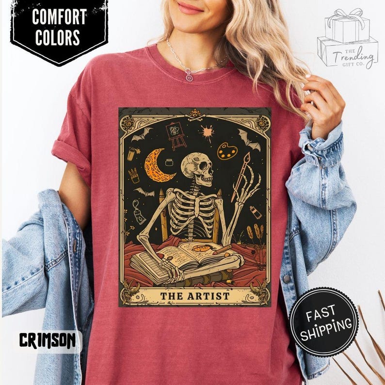 Puede incluir: Camiseta carmes&iacute; con una ilustraci&oacute;n de un artista esqueleto. El dise&ntilde;o incluye una luna creciente, una paleta de pintura y el texto "THE ARTIST". La camiseta tambi&eacute;n tiene las etiquetas "COMFORT COLORS" y "FAST SHIPPING".