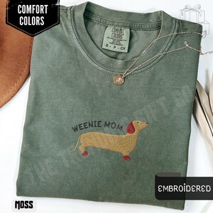 May include: Moss green t-shirt with the words "Weenie Mom" above an embroidered dachshund. The shirt has a crew neck and short sleeves. A gold necklace with a pendant rests on the shirt. The words "Comfort Colors" and "Embroidered" are also visible.