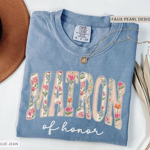 Puede incluir: Camiseta azul vaquero con la palabra "MATRON" en letras florales grandes y "of honor" en escritura. Un collar dorado con un colgante está drapeado sobre la camisa. La camisa tiene una etiqueta Comfort Colors.