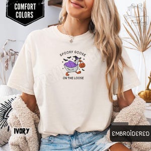 May include: Ivory-colored t-shirt with the text "Spooky Goose on the Loose." The graphic features a goose in a witch hat and cape carrying a jack-o'-lantern. The shirt is embroidered and labeled "Comfort Colors."