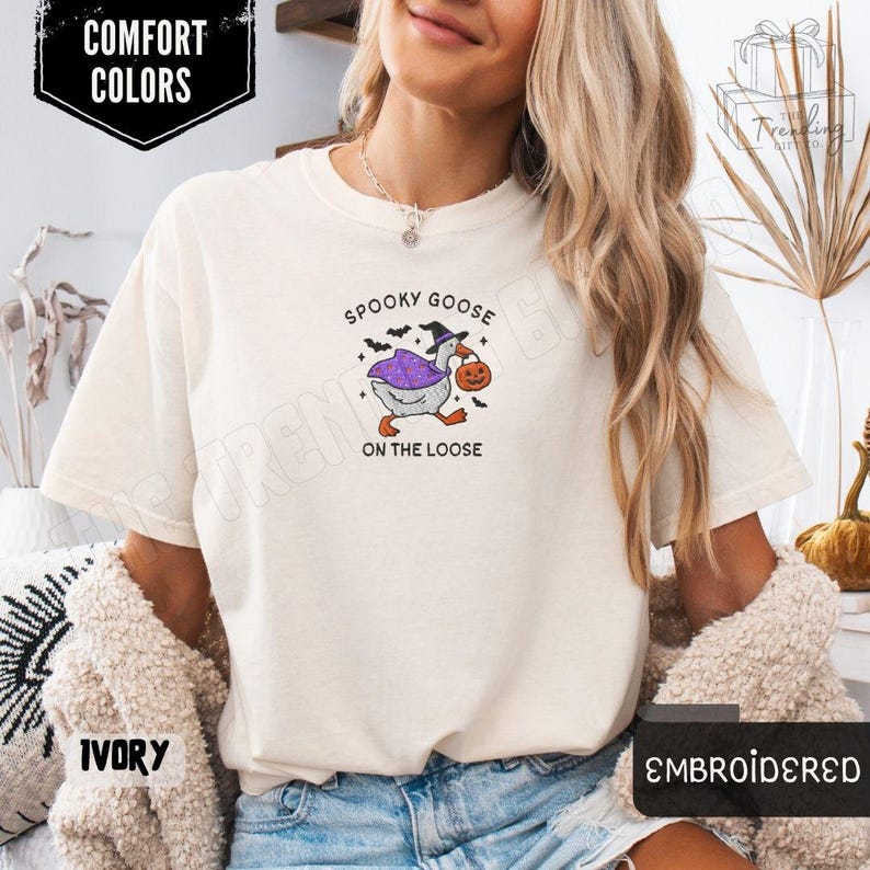 May include: Ivory-colored t-shirt with the text "Spooky Goose on the Loose." The graphic features a goose in a witch hat and cape carrying a jack-o'-lantern. The shirt is embroidered and labeled "Comfort Colors."