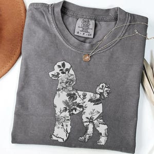 May include: A gray t-shirt with a floral poodle design. The shirt has a distressed look and features the text "Comfort Colors" and "S - P - Ch" on the tag.
