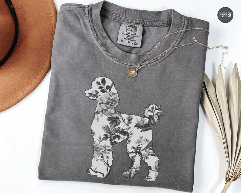 May include: A gray t-shirt with a floral poodle design. The shirt has a distressed look and features the text "Comfort Colors" and "S - P - Ch" on the tag.