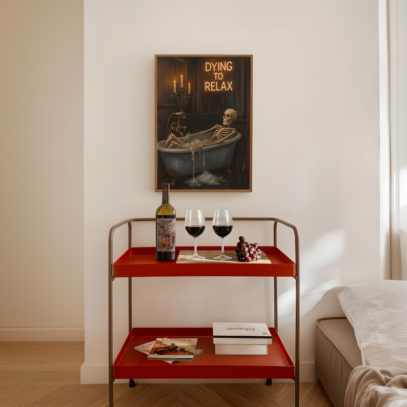 May include: A red serving cart with a bottle of wine, two wine glasses, and a bunch of grapes. A framed artwork depicts a skeleton relaxing in a bathtub with the text "Dying to Relax". The cart has two shelves and a metal frame.