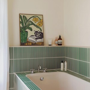 May include: A green and white tiled bathroom with a bathtub and a framed print of a cat reading a book. The print is above a shelf with bottles of toiletries.