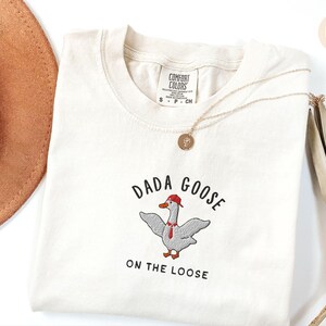 May include: Ivory t-shirt with a goose wearing a red hat and a tie, with the text "Dada Goose on the Loose" embroidered on the front.