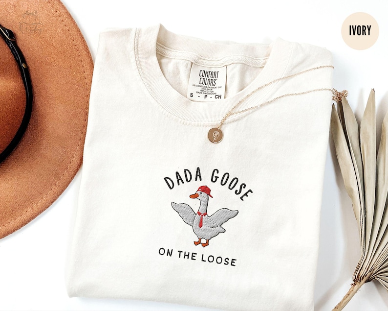 May include: Ivory t-shirt with a goose wearing a red hat and a tie, with the text "Dada Goose on the Loose" embroidered on the front.