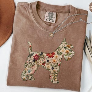 Puede incluir: Una camiseta marr&oacute;n con un dise&ntilde;o bordado floral de un perro Schnauzer. La camiseta tiene el texto "COMFORT COLORS" y "S - P - CH" impreso en la parte delantera.