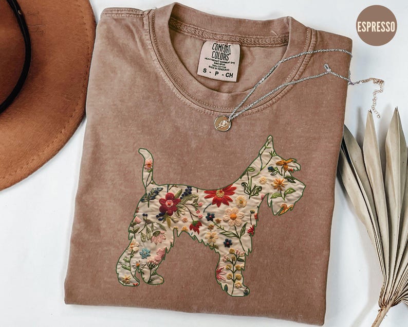 Puede incluir: Una camiseta marr&oacute;n con un dise&ntilde;o bordado floral de un perro Schnauzer. La camiseta tiene el texto "COMFORT COLORS" y "S - P - CH" impreso en la parte delantera.