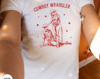 Pode incluir: Camiseta branca com texto vermelho que diz "COWBOY WRANGLER" e uma ilustração vermelha de um cowboy e uma cowgirl.