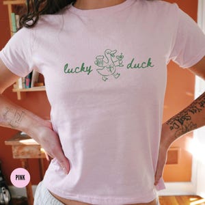 Op de afbeelding: Lichtroze t-shirt met de groene tekst "lucky duck". Een groene cartoon eend is afgebeeld met een bierpul en een cocktail. Het t-shirt heeft een ronde hals en korte mouwen.