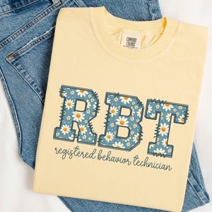 以下が含まれることがあります： 白いデイジー柄の青い文字「RBT」がプリントされた黄色のTシャツ。文字の下には「registered behavior technician」というテキストがプリントされています。