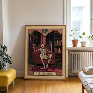 May include: A framed print featuring a skeleton lounging in a burgundy armchair, holding a wine glass. The artwork has a vintage aesthetic, with a dark, ornate background and the text "THE HOMEBODY" at the bottom. The print is displayed in a room with a wooden floor.