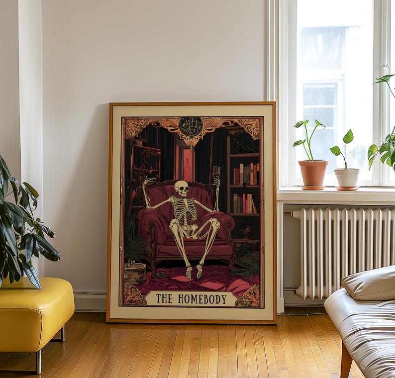 May include: A framed print featuring a skeleton lounging in a burgundy armchair, holding a wine glass. The artwork has a vintage aesthetic, with a dark, ornate background and the text "THE HOMEBODY" at the bottom. The print is displayed in a room with a wooden floor.