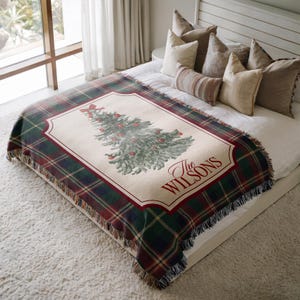 May include: A woven throw blanket featuring a dark green, navy, and red tartan pattern with a central cream-coloured panel. The panel displays a green Christmas tree with red ornaments and a red bow, with the text The WILSONS printed in red.