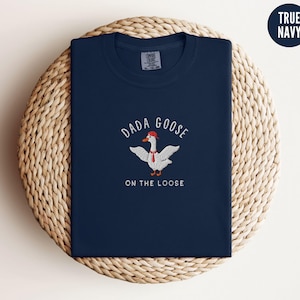 May include: A navy blue t-shirt with a white embroidered goose wearing a red hat and the text "Dada Goose on the Loose".