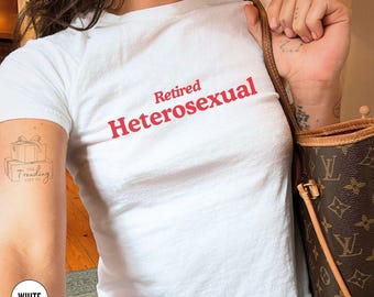 May include: White t-shirt with the text "Retired Heterosexual" in red. The shirt is a crew neck style. The image also shows a brown handbag with a monogram pattern and a tattoo on the arm.