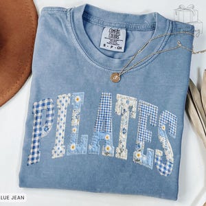 May include: A blue jean Comfort Colors t-shirt with the word "PILATES" appliqued across the front in various blue patterned fabrics and white daisies. A gold necklace with a pendant rests on the shirt.