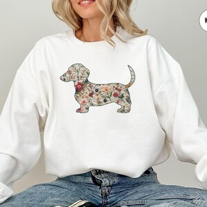 Może przedstawiać: Biała bluza z nadrukiem w kształcie sylwetki psa rasy dachshund ozdobionej kwiatami. Dachshund jest obwiedziony czarną linią i wypełniony kolorowym wzorem kwiatowym.