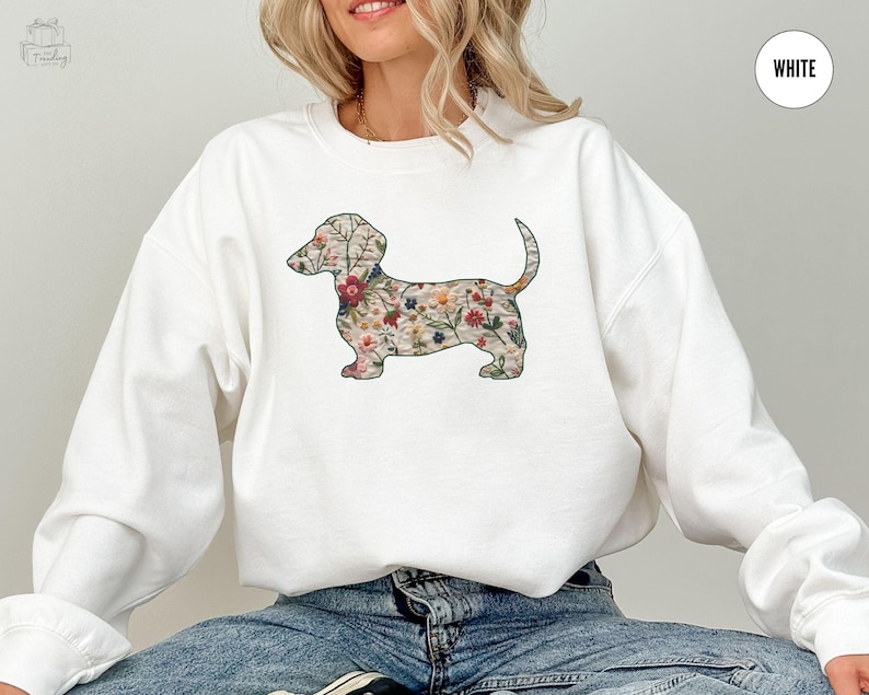 Może przedstawiać: Biała bluza z nadrukiem w kształcie sylwetki psa rasy dachshund ozdobionej kwiatami. Dachshund jest obwiedziony czarną linią i wypełniony kolorowym wzorem kwiatowym.
