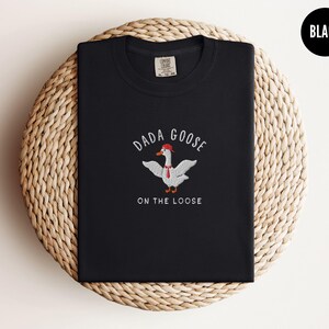 May include: Black t-shirt with a white goose wearing a red hat and a red tie. The text "Dada Goose on the Loose" is embroidered on the shirt.