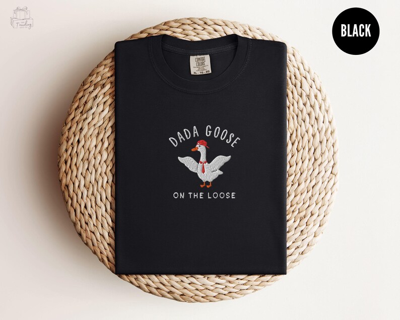 May include: Black t-shirt with a white goose wearing a red hat and a red tie. The text "Dada Goose on the Loose" is embroidered on the shirt.