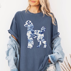 May include: A blue t-shirt with a white floral poodle silhouette design. The shirt is made of a soft, comfortable fabric and has a relaxed fit.