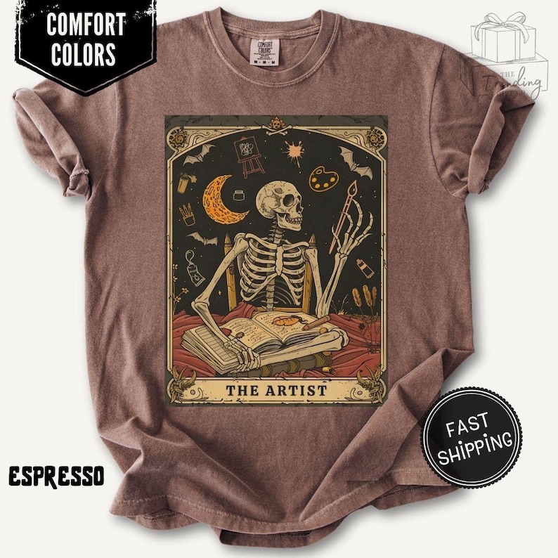 Puede incluir: Camiseta color espresso con un gr&aacute;fico estilo carta del tarot de un artista esqueleto. El dise&ntilde;o incluye un esqueleto sentado en una mesa, sosteniendo un pincel y un libro, con una luna creciente y &uacute;tiles de arte. El texto "THE ARTIST" est&aacute; en la parte inferior.