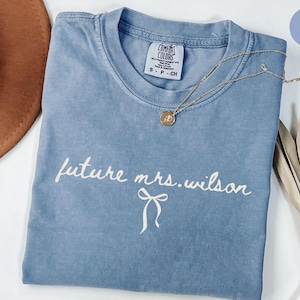 Può includere: Una maglietta di jeans blu con il testo "future mrs. wilson" in scrittura bianca con un fiocco bianco sotto. La maglietta è piegata e appoggiata su una superficie bianca.