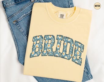 May include: A yellow t-shirt with the word "BRIDE" in blue letters with a white daisy pattern. The letters are in a vintage collegiate style.