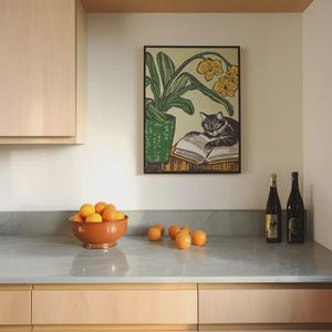 May include: A black cat reading a book while sitting on a table with a potted plant with yellow flowers. The cat is looking at the book. The plant is in a green pot. The table is brown. The background is a light beige wall.