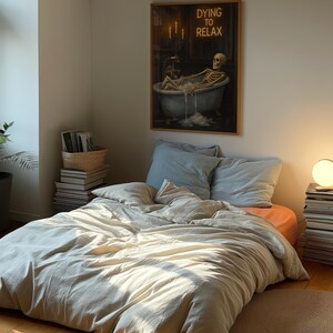 May include: A bedroom scene featuring a bed with light-colored bedding and pillows. A framed artwork on the wall depicts a skeleton relaxing in a bathtub, with the text "DYING TO RELAX". The room is illuminated by natural light.