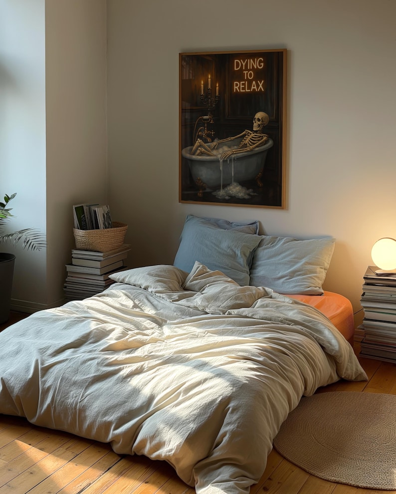 May include: A bedroom scene featuring a bed with light-colored bedding and pillows. A framed artwork on the wall depicts a skeleton relaxing in a bathtub, with the text "DYING TO RELAX". The room is illuminated by natural light.