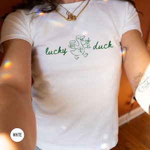 Op de afbeelding: Wit T-shirt met een groene afbeelding van een eend met een bierpul en een cocktail, met een klavertje vier. De woorden "lucky duck" staan in groene letters boven de eend. Het shirt is gemaakt van een zacht materiaal.