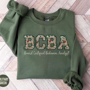 Peut inclure: Un sweat-shirt vert avec le texte "BCBA Board Certified Behavior Analyst" en lettres blanches avec un motif floral.