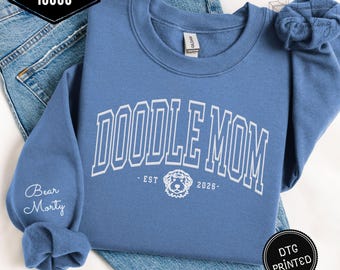 Puede incluir: Sudadera azul índigo con el texto blanco "DOODLE MOM" arqueado sobre el pecho. Debajo, "EST 2026" y un gráfico de perro doodle. La manga izquierda tiene el texto "Bear Morty". La sudadera está doblada sobre unos vaqueros azules.