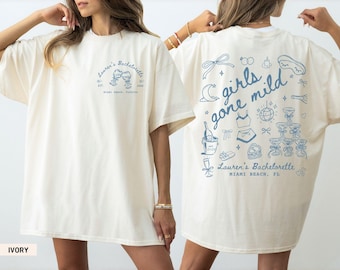 Op de afbeelding: Ivoor kleurig t-shirt met blauwe tekst en afbeeldingen. De voorkant toont "Lauren's Bachelorette" met wijnglazen. De achterkant toont "girls gone mild" met illustraties met feestthema. "Miami Beach, FL" is ook gedrukt.