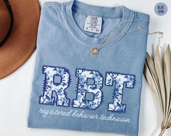 Könnte beinhalten: Ein hellblaues T-Shirt mit einem floralen Design, das "RBT" buchstabiert, und dem Text "registered behavior technician".