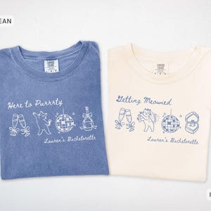 Puede incluir: Dos camisetas, una azul y otra marfil, con diseños gráficos blancos. La camiseta azul dice "Here to Purrrty" con ilustraciones de gato y bola de discoteca. La camiseta marfil dice "Getting Meowied" con gráficos similares. Ambas camisetas dicen "Lauren's Bachelorette".