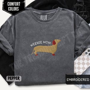 May include: Pepper-colored long-sleeve shirt with the text "WEENIE MOM" and an embroidered dachshund. The dachshund is tan with a red head and feet. The shirt is part of the Comfort Colors collection and is embroidered.