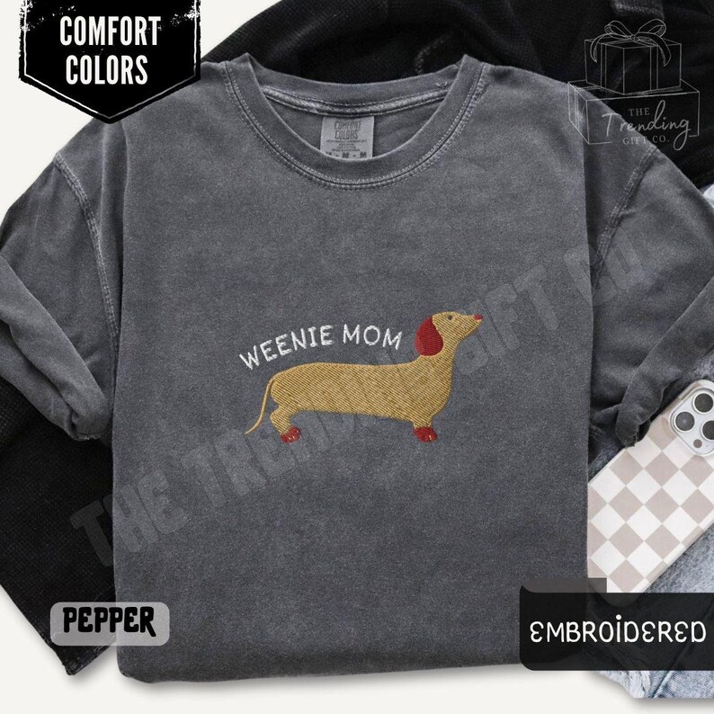 May include: Pepper-colored long-sleeve shirt with the text "WEENIE MOM" and an embroidered dachshund. The dachshund is tan with a red head and feet. The shirt is part of the Comfort Colors collection and is embroidered.
