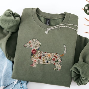 Może przedstawiać: Zielona bluza z nadrukiem kwiatowego jamnika. Bluza wykonana jest z miękkiego, wygodnego materiału i ma luźny kr&oacute;j.